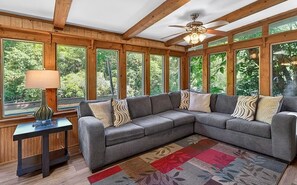 Ein Zimmer mit einer großen Eckcouch, einem kleinen Tisch mit Lampe und großen Fenstern mit Blick auf Grünflächen.