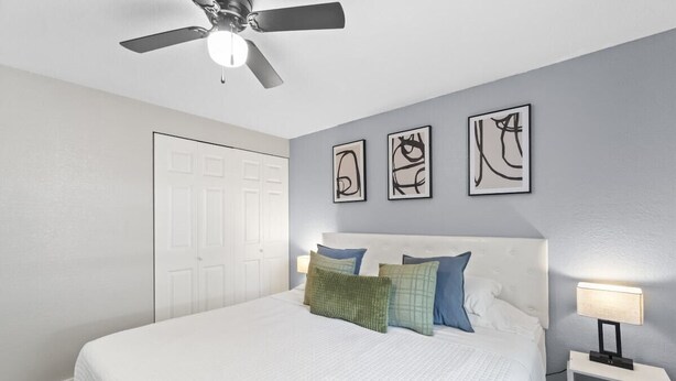A bedroom with a bed, a ceiling fan, a nightstand with a lamp, and abstract wall art.