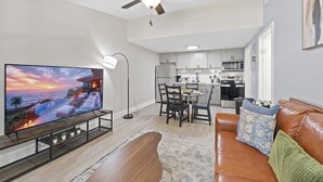 A living room with a television, a ceiling fan, a dining area with chairs, and a kitchen in the background.