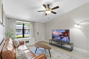 Een woonkamer met een leren bank, een televisie op een standaard, een plafondventilator en een raam met gordijnen.