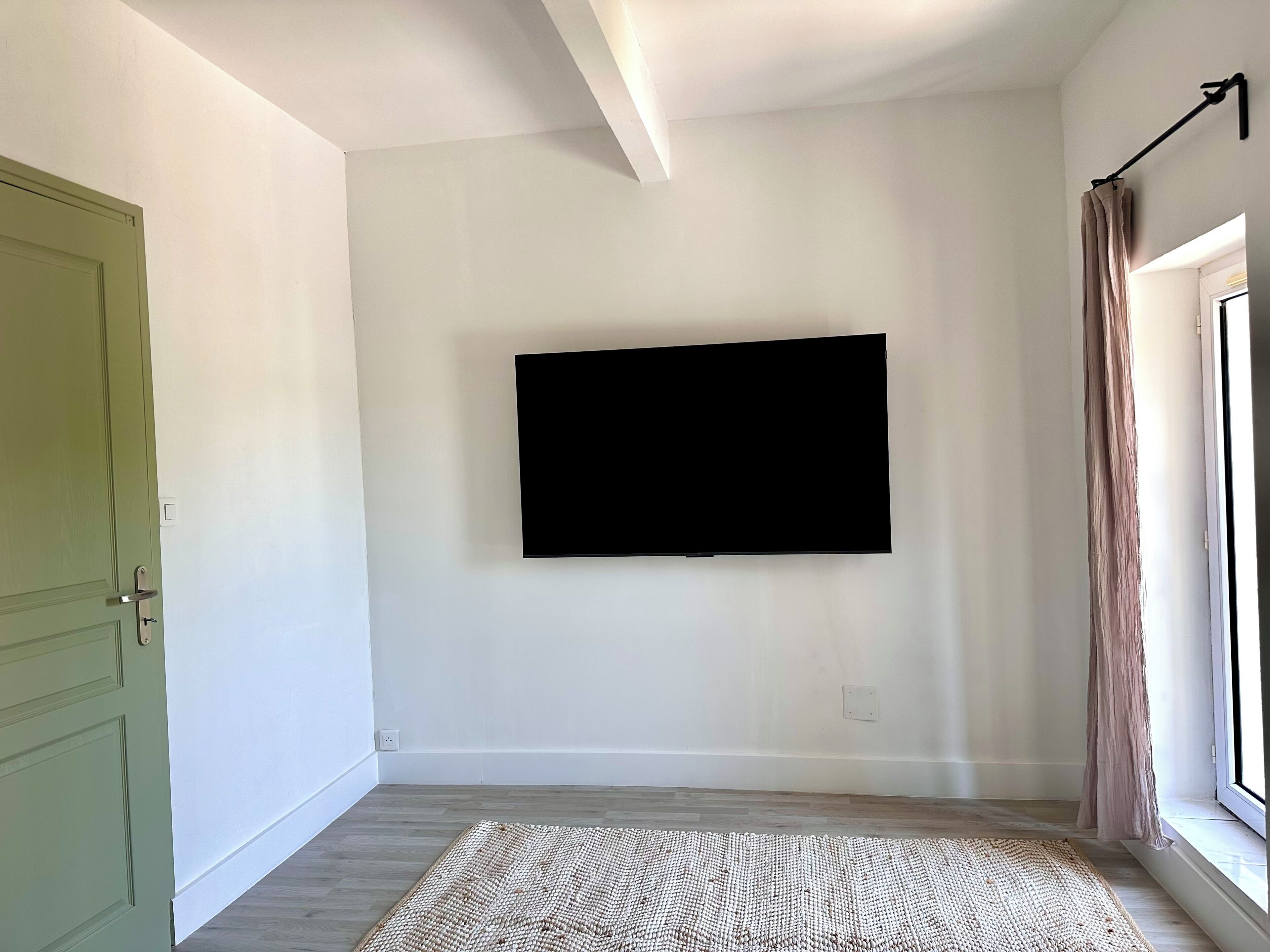 Une pièce avec un téléviseur fixé au mur, une porte verte, une fenêtre avec des rideaux et un tapis tissé sur le sol.
