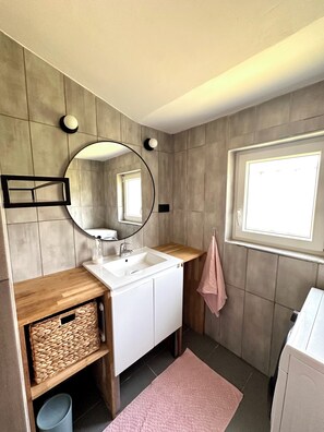 A bathroom with a white sink, wooden vanity, a round mirror, a towel rack, and a window. - Family home between Avignon and Mont Ventoux with free EV charging (Pernes-les-Fontaines)