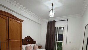 Un dormitorio con cama, armario de madera, una mesita, una silla, un espejo y una ventana con cortinas.