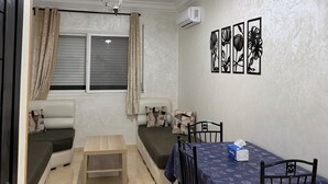 Una habitación con una mesa de comedor, sillas, un sofá, una ventana con cortinas, una lámpara de techo y decoración en las paredes.