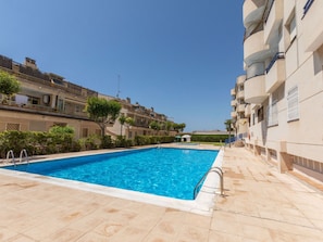 Una piscina con agua cristalina azul, rodeada por un solárium con baldosas, y edificios de varios pisos con balcones adyacentes.