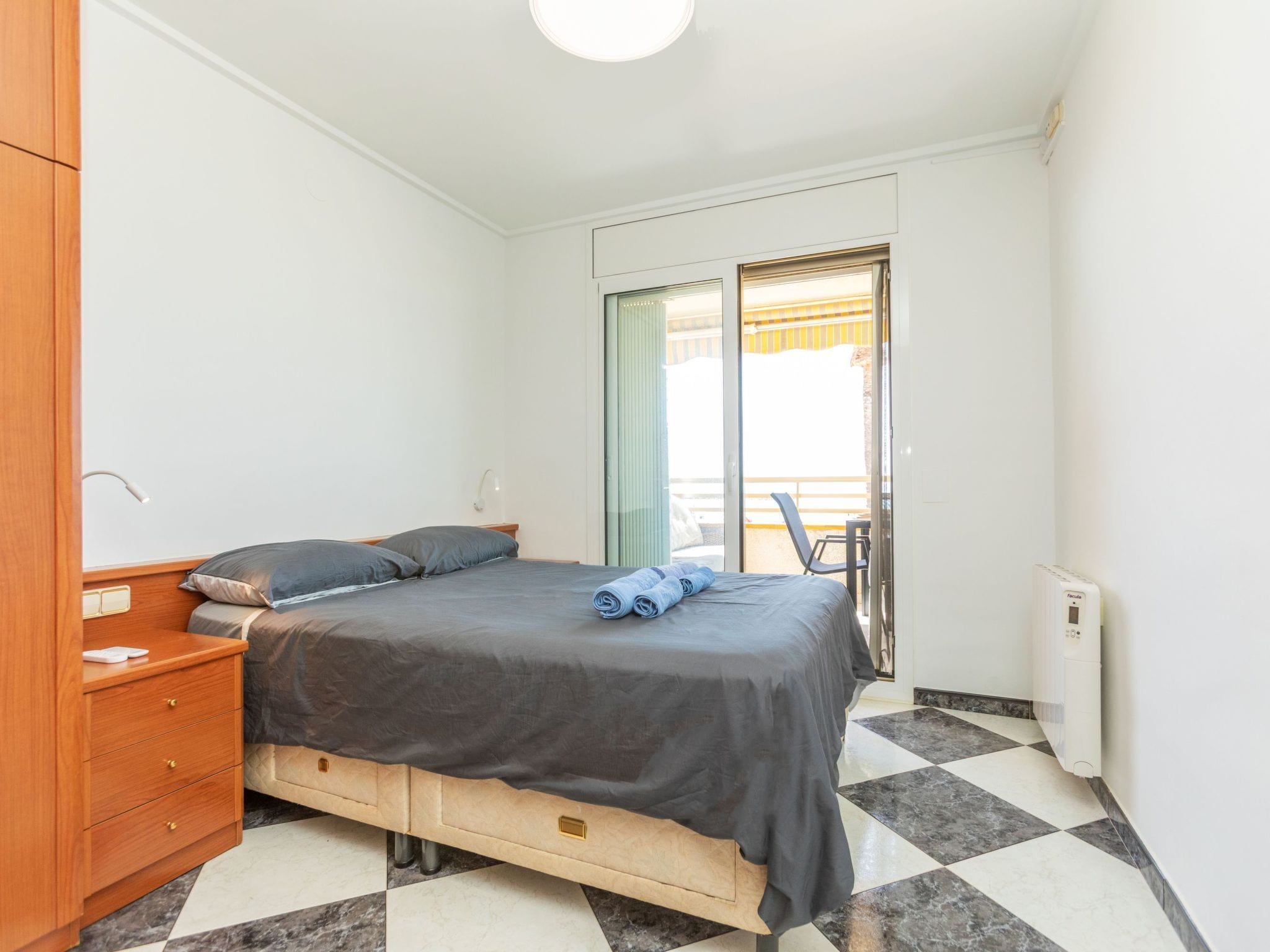Ein Schlafzimmer mit Bett, einem hölzernen Nachttisch, einem Schreibtisch, einem Stuhl und Blick auf den Balkon.