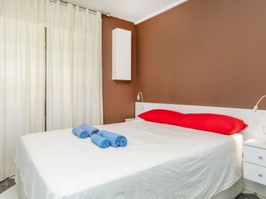 Ein Hotelzimmer mit Bett, weißen Vorhängen, einem roten Kopfkissen und einem weißen Nachttisch.