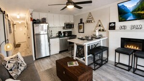 Uma cozinha moderna com armários brancos, uma área de jantar com mesa branca e banquetas pretas, uma lareira e uma TV fixada na parede.