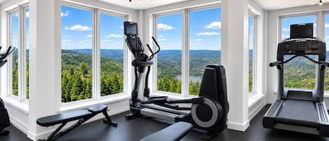 Un gimnasio con equipo de ejercicio, ventanas amplias y vista a un bosque.