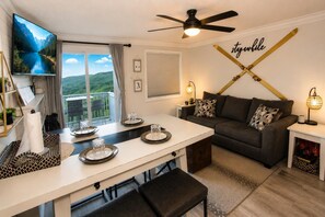 A modern living room with a dining area, a ceiling fan, a TV, and a view of a mountain landscape. - ML 290: Mountain Bliss at Snowshoe | Summer Trails & Breathtaking Views (Snowshoe)