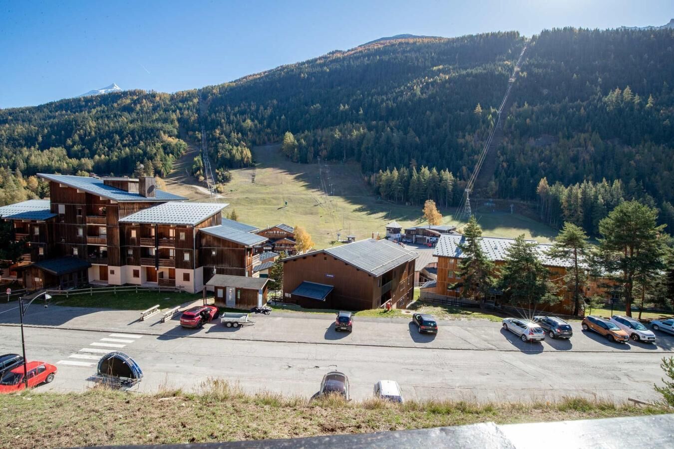 Un chalet de montagne comprenant plusieurs bâtiments, un parking et offrant une vue sur une vallée boisée.