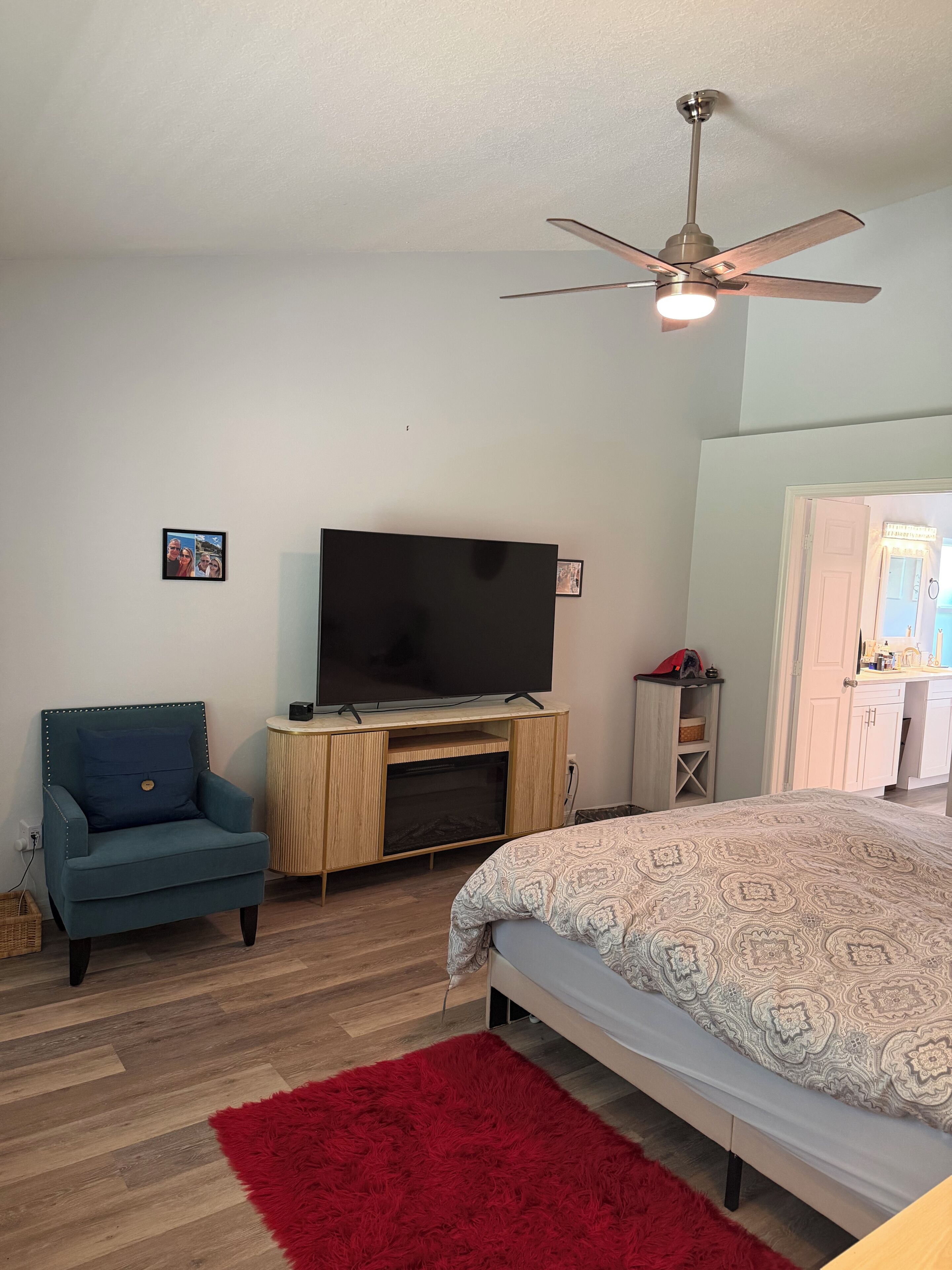Un dormitorio con una cama, un televisor sobre un mueble de madera, un sillón azul, un tapete rojo y un ventilador de techo.