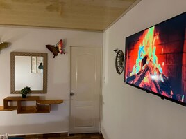 Una habitación con una puerta blanca, una gran pantalla de televisión mostrando un fuego, un banco de madera y una decoración de mariposa.