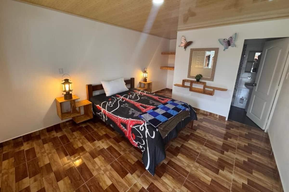 A bedroom with a wooden floor, a bed with a patterned bedspread, bedside lamps, a mirror, and a butterfly decoration on the wall.