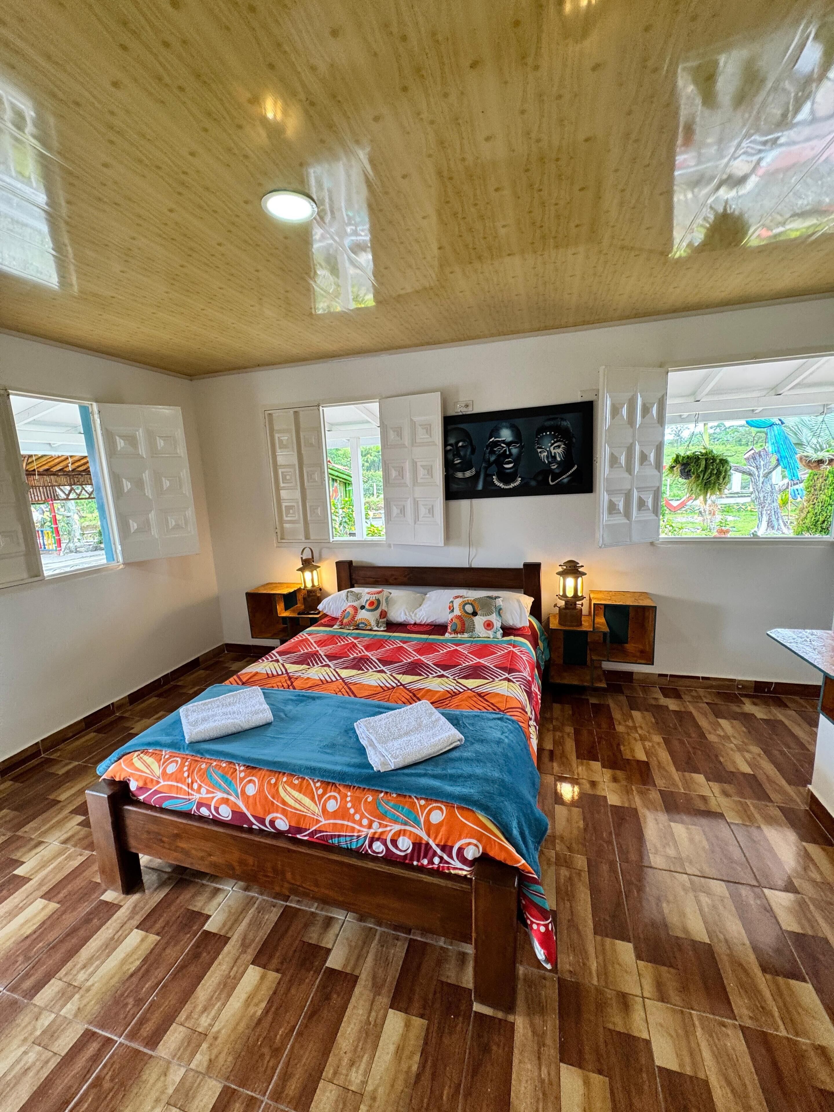 Um quarto com piso de madeira, uma cama com roupa de cama colorida, duas mesas de cabeceira com luminárias e uma janela grande com vista para o verde.
