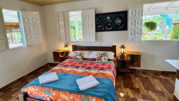 Um quarto com piso de madeira, uma cama com roupa de cama colorida, duas mesas de cabeceira com luminárias e uma janela grande com vista para o verde.