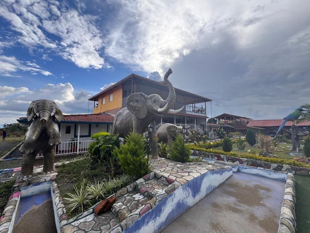 Un edificio con techo rojo y balcón, rodeado de un jardín ajardinado con un pequeño estanque y estatuas.