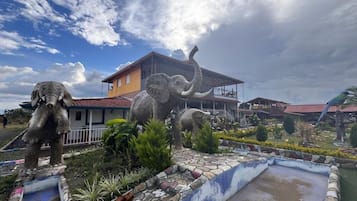 Un edificio con techo rojo y balcón, rodeado de un jardín ajardinado con un pequeño estanque y estatuas.