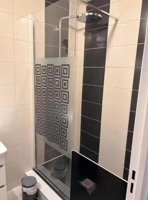 Um chuveiro com paredes de vidro, um padrão de azulejos quadrados na parede, uma pia branca e uma lixeira.