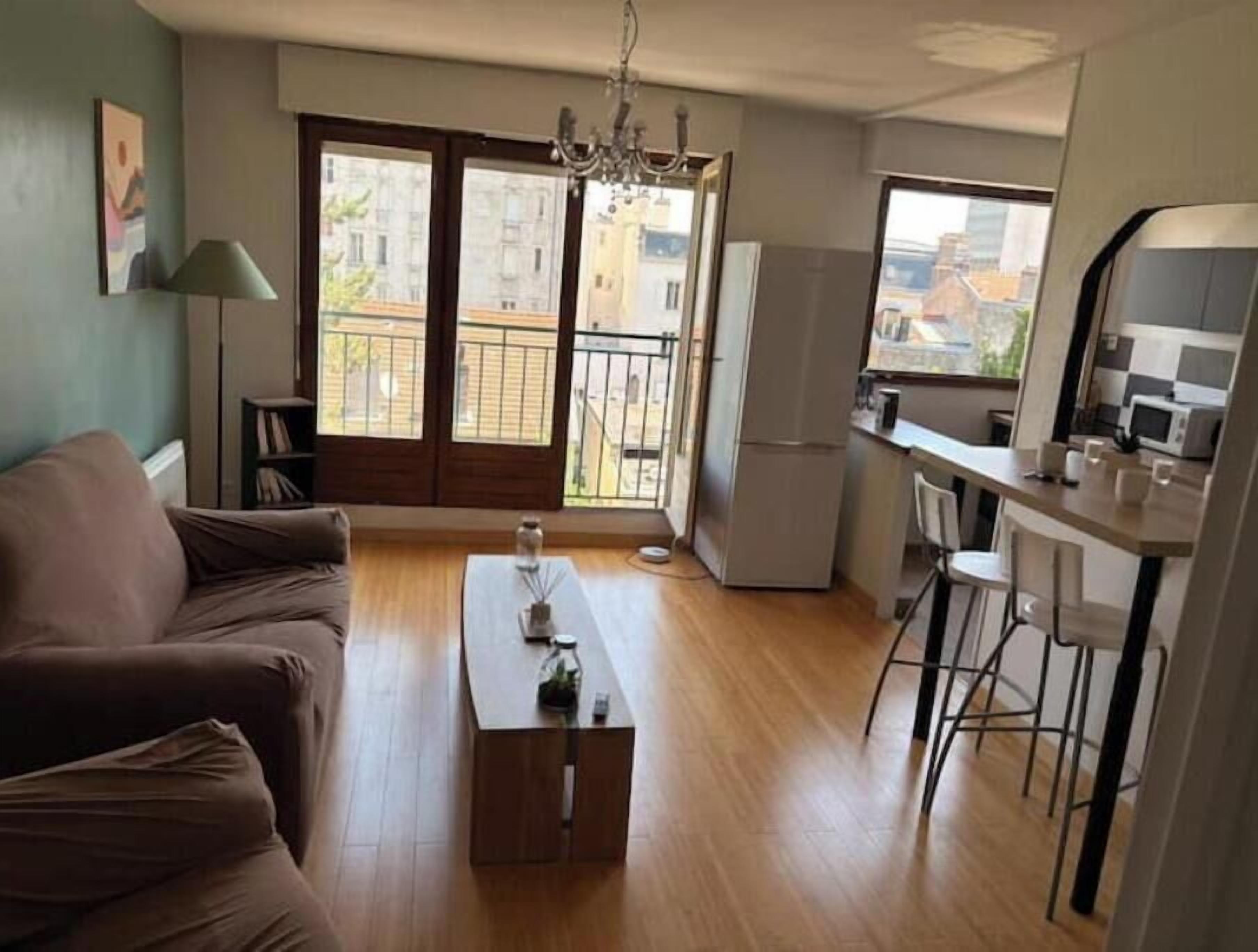 A living room with a sofa, a coffee table, a dining area with chairs, a kitchen with a refrigerator, and a balcony with a view of buildings.