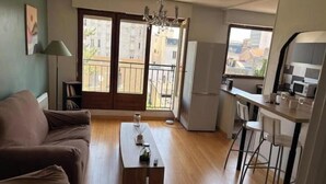 A living room with a sofa, a coffee table, a dining area with chairs, a kitchen with a refrigerator, and a balcony with a view of buildings. - Apartment 5 minutes from the Nancy train station (Nancy)