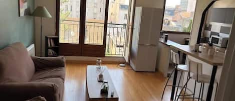 A living room with a sofa, a coffee table, a dining area with chairs, a kitchen with a refrigerator, and a balcony with a view of buildings.