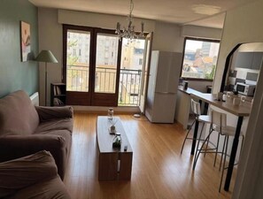 A living room with a sofa, a coffee table, a dining area with chairs, a kitchen with a refrigerator, and a balcony with a view of buildings. - Apartment 5 minutes from the Nancy train station (Nancy)