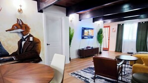 Una stanza con pavimento in legno, un tavolo rotondo, sedie, un divano, una televisione e una pianta fissata al muro.