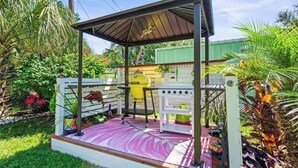 Een overdekt terras buiten met een barbecue, een witte schutting en diverse planten.