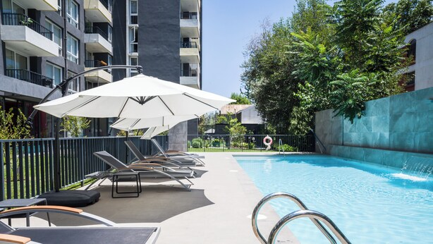 Piscina con sillas de descanso y sombrillas, rodeada por una cerca y follaje.