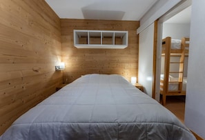 A bedroom with a wooden wall, a bed with a white duvet, a bedside table with a lamp, and a bunk bed in the background. - 20m from ski lifts, 3rd floor, swimming pool, sauna, balcony, tv, ski locker, 33m², Les Orres (Les Orres)
