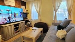 Ett vardagsrum med en tv-skärm som visar en strandscen, en soffa, ett soffbord och ett litet skrivbord med en stol.
