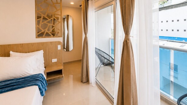 A hotel room with a bed, a wooden headboard, a mirror, a window with curtains, and a view of a building outside. -  Hotel Azure Cartagena (Cartagena)