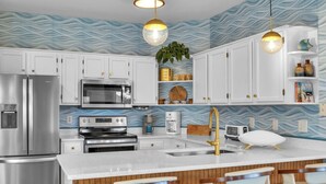 A modern kitchen with white cabinets, stainless steel appliances, a central island with bar stools, and a wavy blue backsplash. - Sea Trail Condo with Golf, Pools, Tennis & Beach Access (Sunset Beach)
