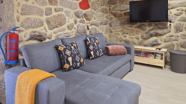A stone wall, grey sofa, patterned cushions, a flat-screen TV, and a fire extinguisher. - Coração de Dorna House | Castro Laboreiro | Peneda-Gerês National Park (Castro Laboreiro)