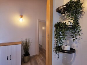 Ein Flur mit Holzboden, einer weißen Tür, einer Wandleuchte, einem Blumentopf und einem Regal mit einer hängenden Pflanze und einer Toilettenpapierrolle.