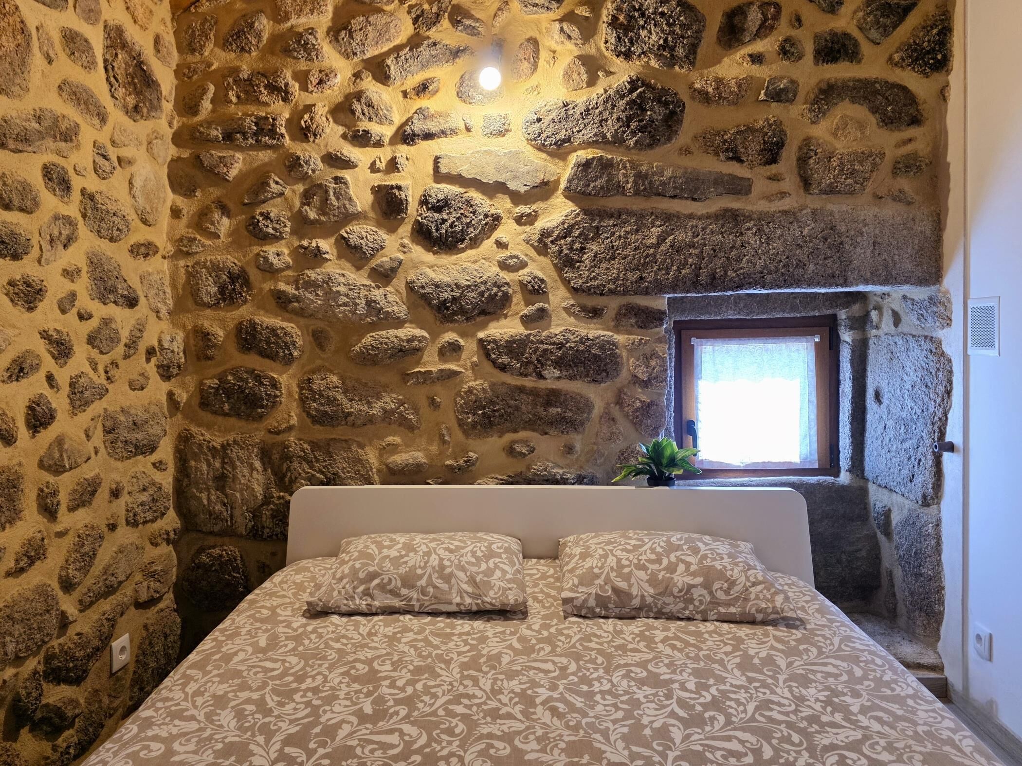 Un dormitorio con pared de piedra, cama con ropa de cama estampada y ventana con vistas.