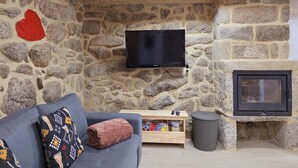 A stone-walled room with a television mounted on the wall, a sofa with patterned cushions, a small wooden table with remote controls, and a fireplace. - Coração de Dorna House | Castro Laboreiro | Peneda-Gerês National Park (Castro Laboreiro)
