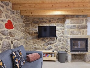 A stone-walled room with a television mounted on the wall, a sofa with patterned cushions, a small wooden table with remote controls, and a fireplace. - Coração de Dorna House | Castro Laboreiro | Peneda-Gerês National Park (Castro Laboreiro)