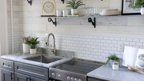 Una cocina con lavamanos, estufa y armarios, con azulejos tipo metro blancos, estanterías de metal negro y una alfombra morada.