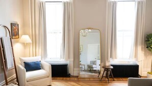 Un salón luminoso con un sofá blanco, una almohada azul, piso de madera y un espejo grande.