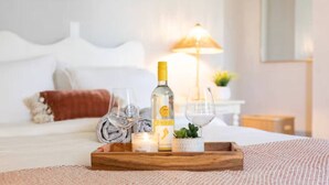 Una cama bien hecha con sábanas blancas y una almohada roja, una mesita de noche con una lámpara, y una bandeja con una botella, vasos y una vela.