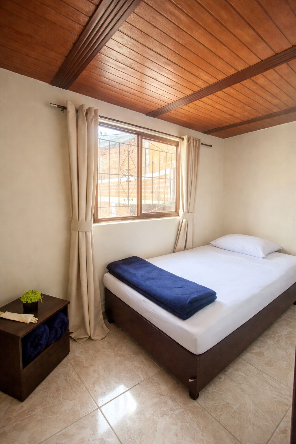 A bedroom with a bed, a window with curtains, a bedside table with a plant, and a wooden ceiling.