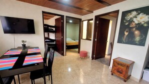 Un coin repas avec une table et des chaises, un téléviseur fixé au mur et une chambre à coucher visible à travers une porte ouverte.
