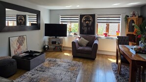 A living room with a flat-screen TV, a leather armchair, a dining table, and a large mirror. - Spacious 2 bedroom maisonette (Broughton)