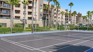 Un campo da tennis con una rete, circondato da una recinzione e palme, con un edificio a più piani sullo sfondo.