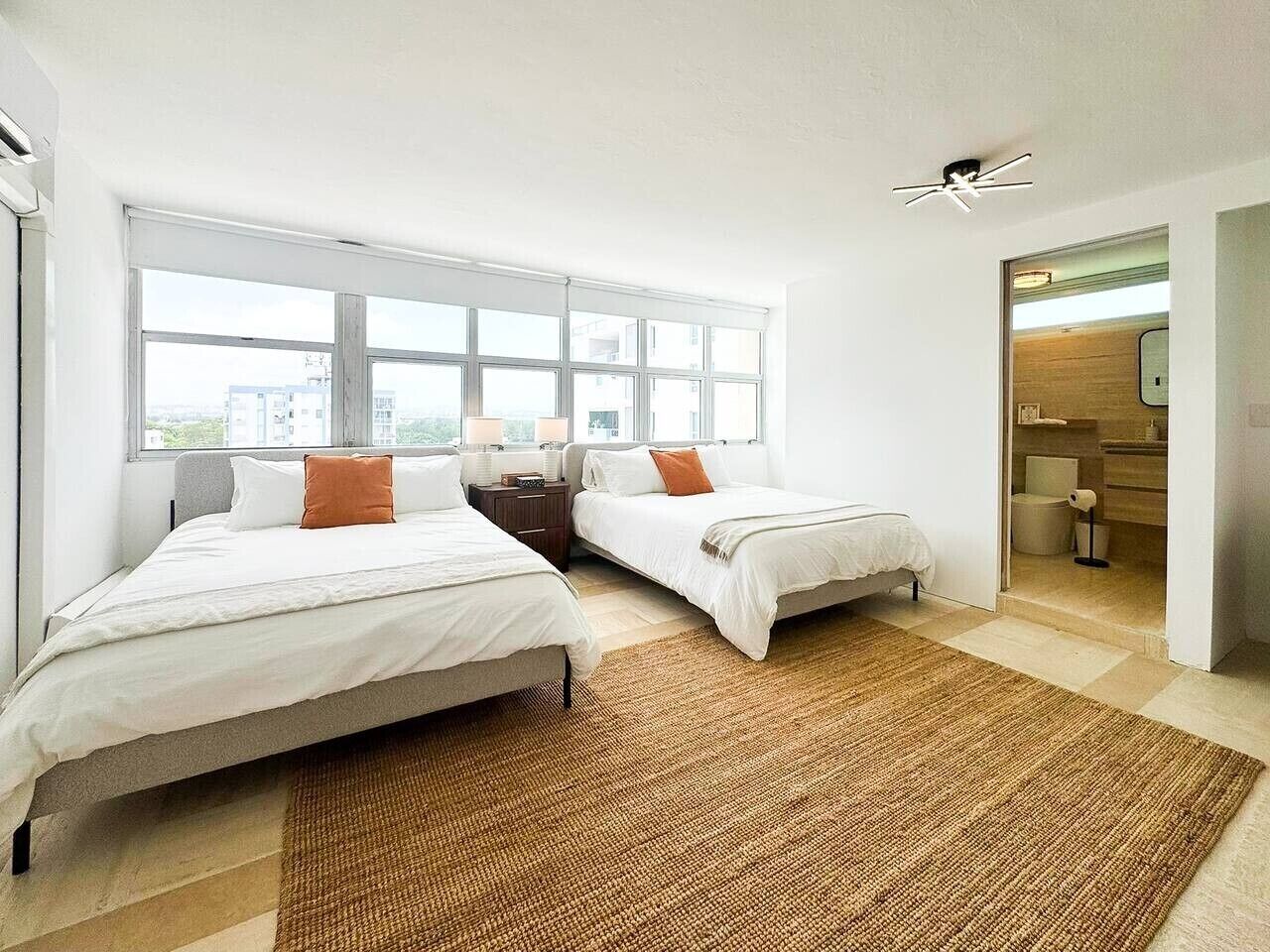 A modern hotel room with two beds, a large window, a ceiling fan, and a visible bathroom.