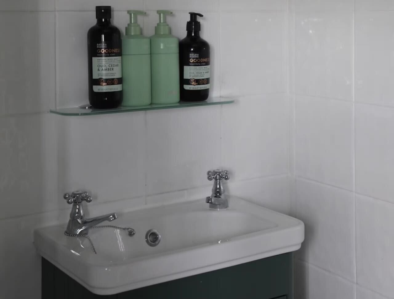Une salle de bain avec un lavabo blanc, deux robinets et trois bouteilles de gel douche de la marque Goodness, posées sur une étagère au-dessus du lavabo.