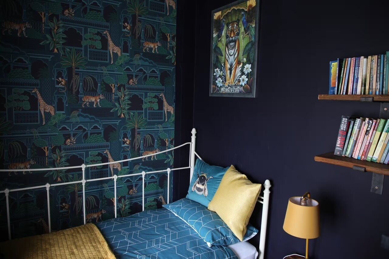 A bed with a white metal frame, a blue patterned bedspread, and a yellow pillow. There is a wall with a jungle-themed wallpaper and a framed picture above the bed. A bookshelf with books is to the right of the bed.