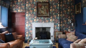 Une pièce avec une cheminée, un papier peint à motifs floraux, un canapé en cuir marron et un fauteuil bleu.